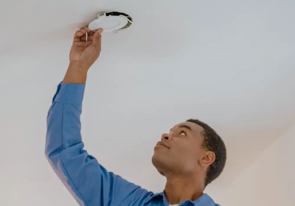 Electrician with short hair wearing a blue work shirt, installing or checking a circular LED light fixture in a white ceiling.