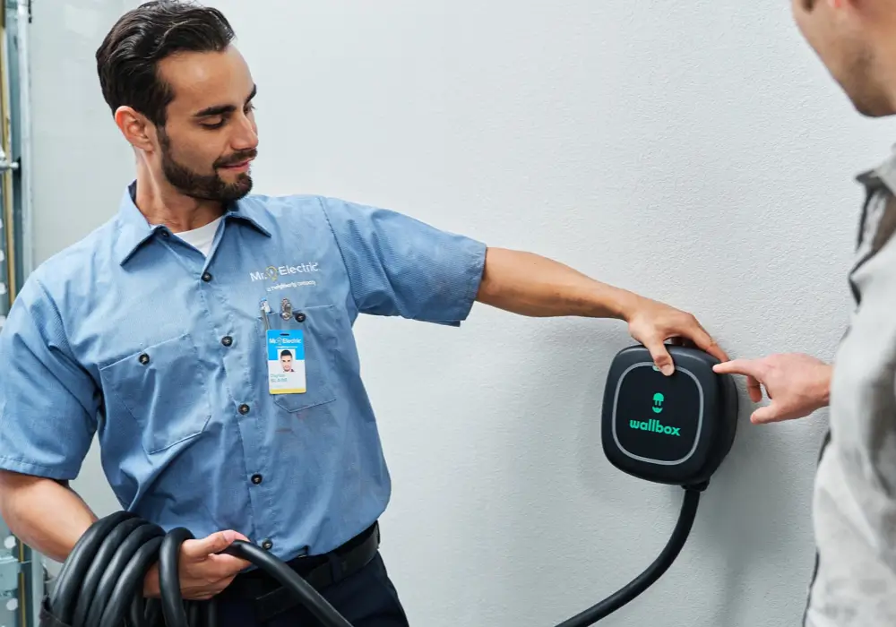 Smiling service professional in a blue striped shirt with company badge demonstrating a black Wallbox electric vehicle charger, holding the charging cable, gesturing toward a customer, against a white indoor-outdoor wall.