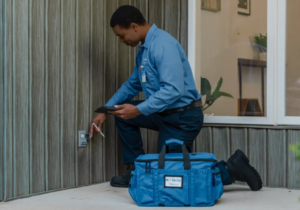 Service professional wearing a blue work shirt and dark pants kneeling outdoors, examining an exterior wall outlet with a pen and clipboard beside a blue tool bag, next to a modern building facade under a window.