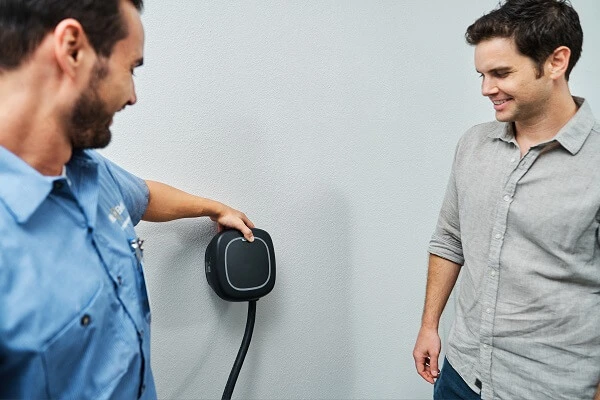 A smiling Mr. Electric electrician touches an EV charger on the wall to show another smiling man watching how to use it.